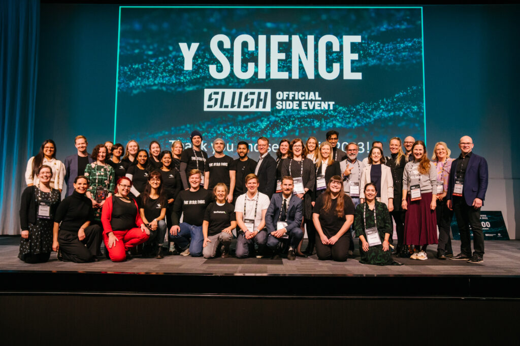 Stage with all Y Science event organisers, volunteers, panelists and speakers, as well as people from the pitching competition. Text behind the stage reads Y Science: Slush Official Side Event in white text over a textured teal background. 